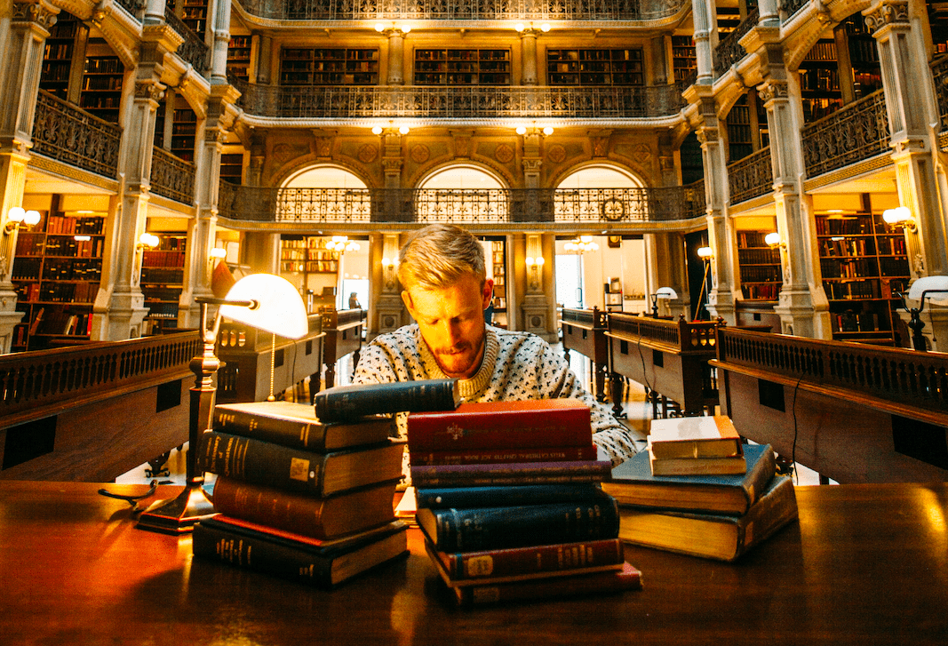 Ein Mann sitzt an einem Holztisch in einer alten Bibliothek und lernt Englische Grammatik von vielen Büchern.